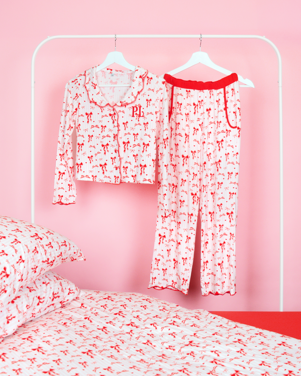 Girls P.Louise Christmas bow pyjama trousers set featuring an all over red bow design against pink and white stripes hanging on coat hangers in a pink bedroom with matching bedding