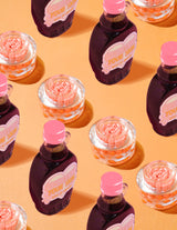 Διαφανή δοχεία P.Louise Dose of Dreams Vitamin C Capsule Cream με πορτοκαλί κάψουλες μέσα τους που περιβάλλονται από μωβ και ροζ μπουκάλια σιρόπι σε πορτοκαλί φόντο
