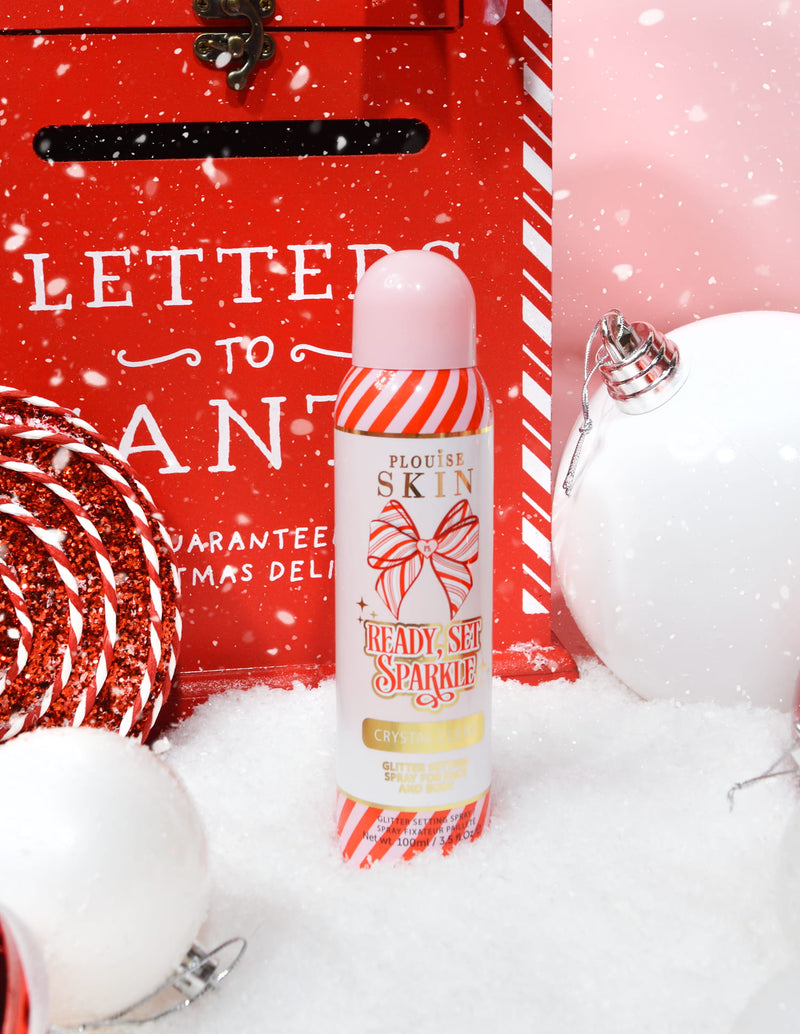 Ένα μπουκάλι P. Louise Ready Set Sparkle Christmas Glitter Setting Spray που περιβάλλεται από εορταστικές διακοσμήσεις όπως χιόνι, μπιχλιμπίδια και ένα χριστουγεννιάτικο γραμματοκιβώτιο